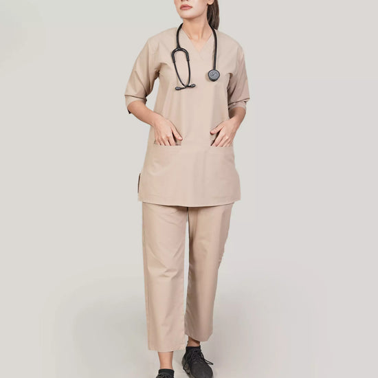 A woman wearing a beige scrub set with a V-neckline design, 3-quarter sleeves, and two bone pockets. She is also wearing matching beige trousers with a pajama-style fit and two pockets.