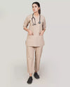 A woman wearing a beige scrub set with a V-neckline design, 3-quarter sleeves, and two bone pockets. She is also wearing matching beige trousers with a pajama-style fit and two pockets.