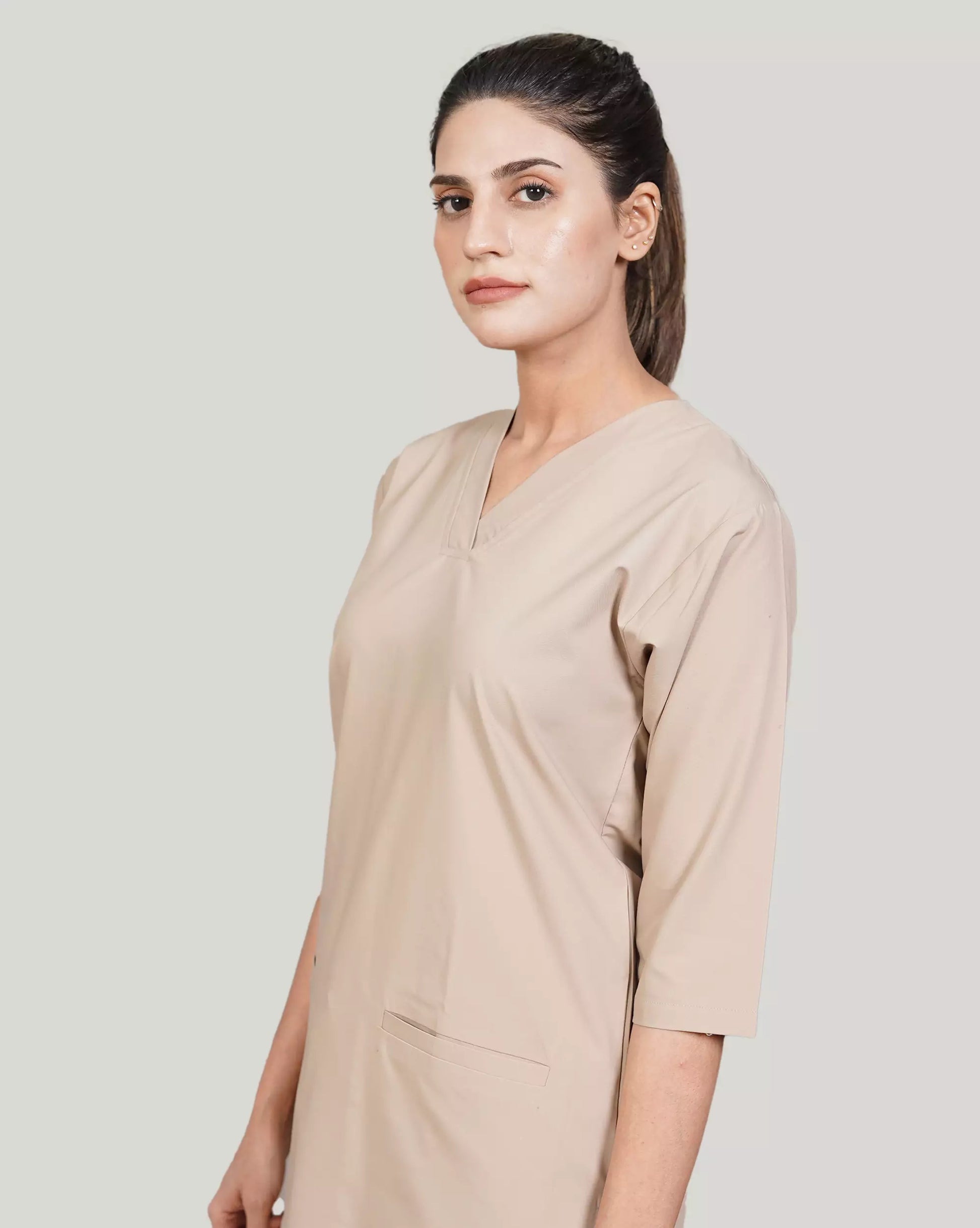 A woman wearing a green scrub set with a V-neckline and 3-quarter sleeves, featuring two bone pockets on the shirt and two pockets on the trousers. She is smiling and standing against a white background.