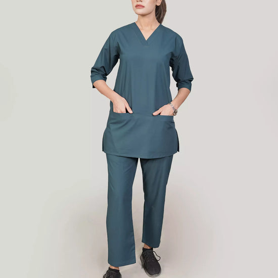 A woman wearing a green scrub set with a V-neckline and 3-quarter sleeves, featuring two bone pockets on the shirt and two pockets on the trousers. She is smiling and standing against a white background.