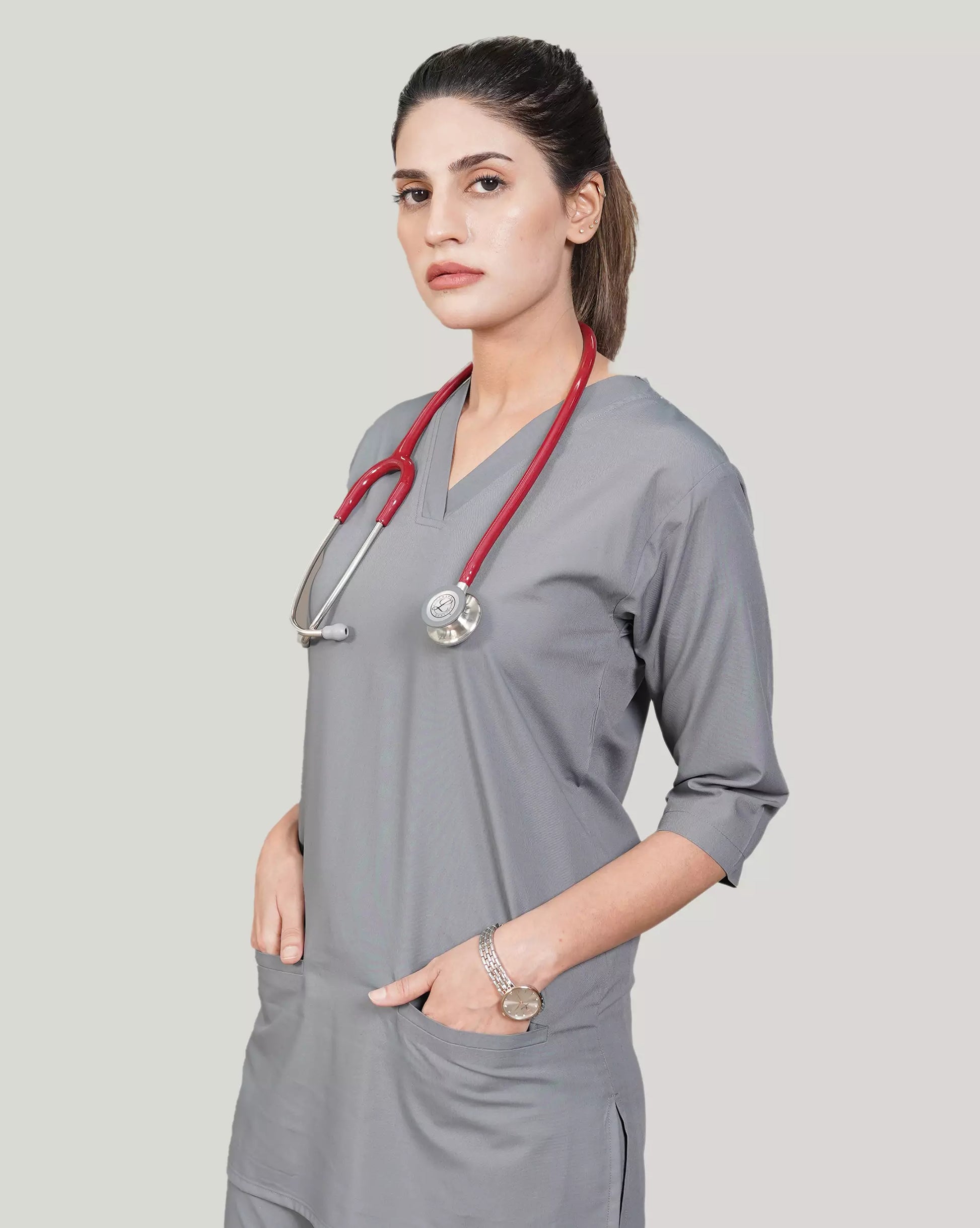 A woman wearing a green scrub set with a V-neckline and 3-quarter sleeves, featuring two bone pockets on the shirt and two pockets on the trousers. She is smiling and standing against a white background.