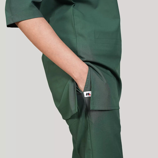 A woman wearing a green scrub set with a V-neckline and 3-quarter sleeves, featuring two bone pockets on the shirt and two pockets on the trousers. She is smiling and standing against a white background.
