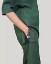 A woman wearing a green scrub set with a V-neckline and 3-quarter sleeves, featuring two bone pockets on the shirt and two pockets on the trousers. She is smiling and standing against a white background.