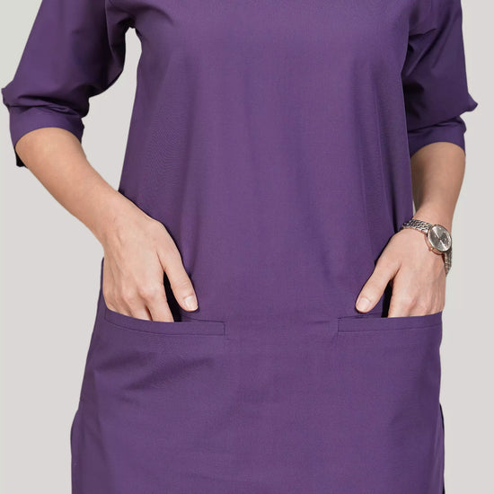 A woman wearing a green scrub set with a V-neckline and 3-quarter sleeves, featuring two bone pockets on the shirt and two pockets on the trousers. She is smiling and standing against a white background.