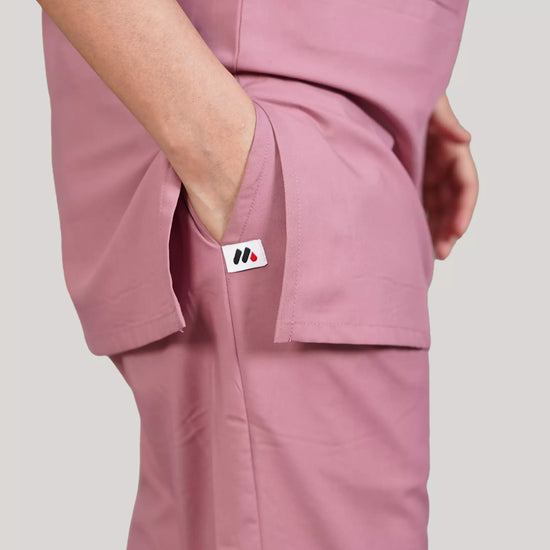 A woman wearing a green scrub set with a V-neckline and 3-quarter sleeves, featuring two bone pockets on the shirt and two pockets on the trousers. She is smiling and standing against a white background.