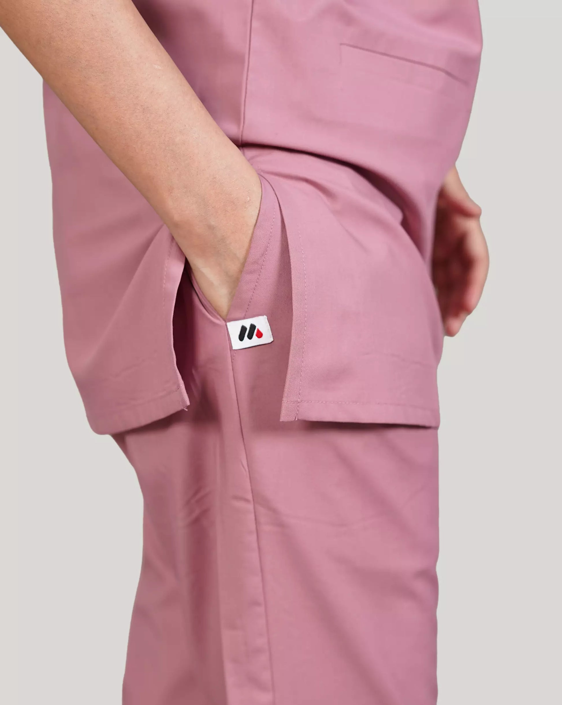 A woman wearing a green scrub set with a V-neckline and 3-quarter sleeves, featuring two bone pockets on the shirt and two pockets on the trousers. She is smiling and standing against a white background.