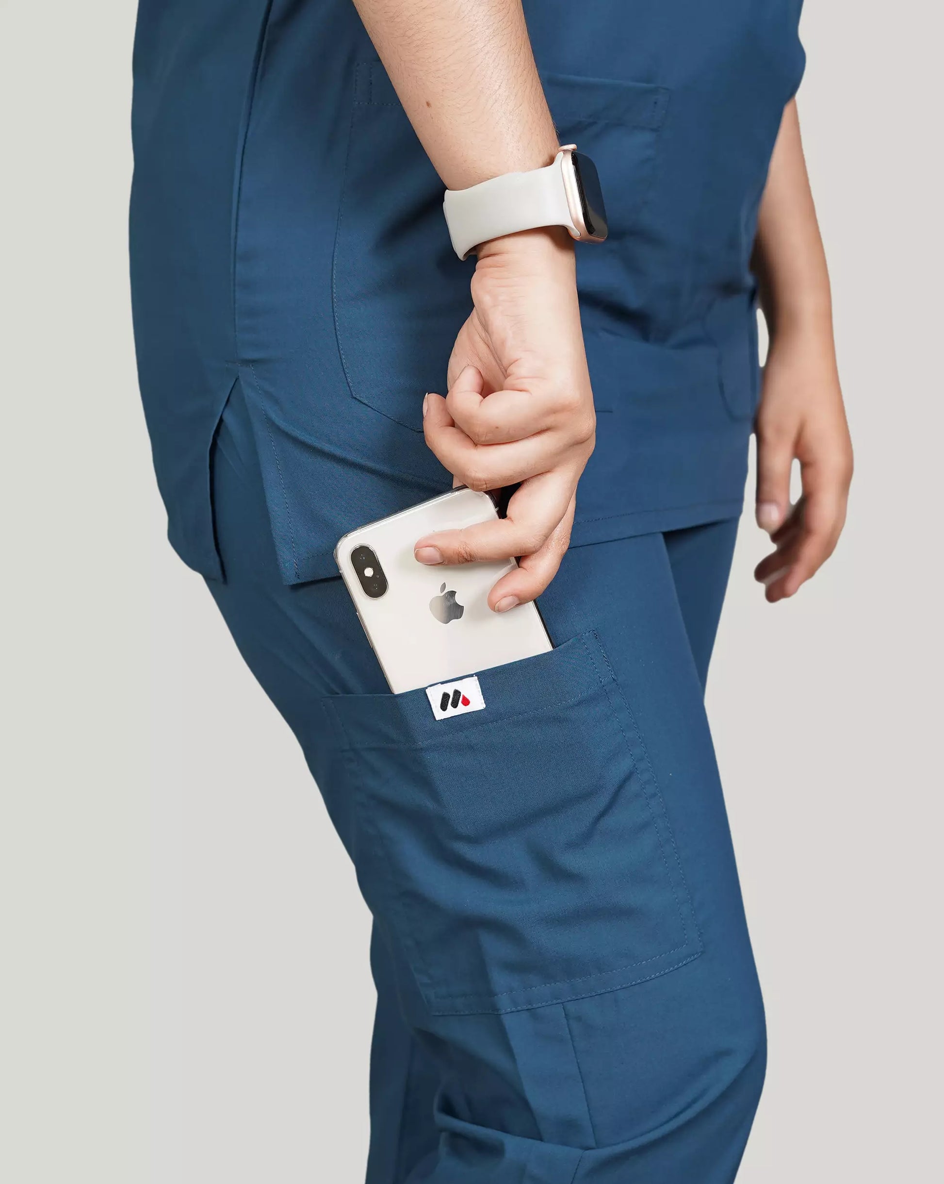 Close-up of a hand inserting a smartphone into the pocket of blue 'Zinc' Medfit unisex scrub pants, featuring the Medfit logo and smartwatch for convenient access.