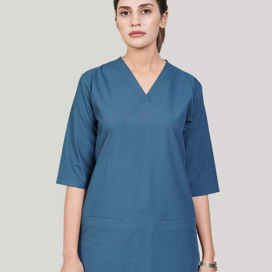 A woman wearing a green scrub set with a V-neckline and 3-quarter sleeves, featuring two bone pockets on the shirt and two pockets on the trousers. She is smiling and standing against a white background.