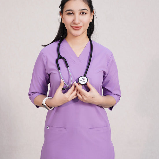 A woman wearing a green scrub set with a V-neckline and 3-quarter sleeves, featuring two bone pockets on the shirt and two pockets on the trousers. She is smiling and standing against a white background.
