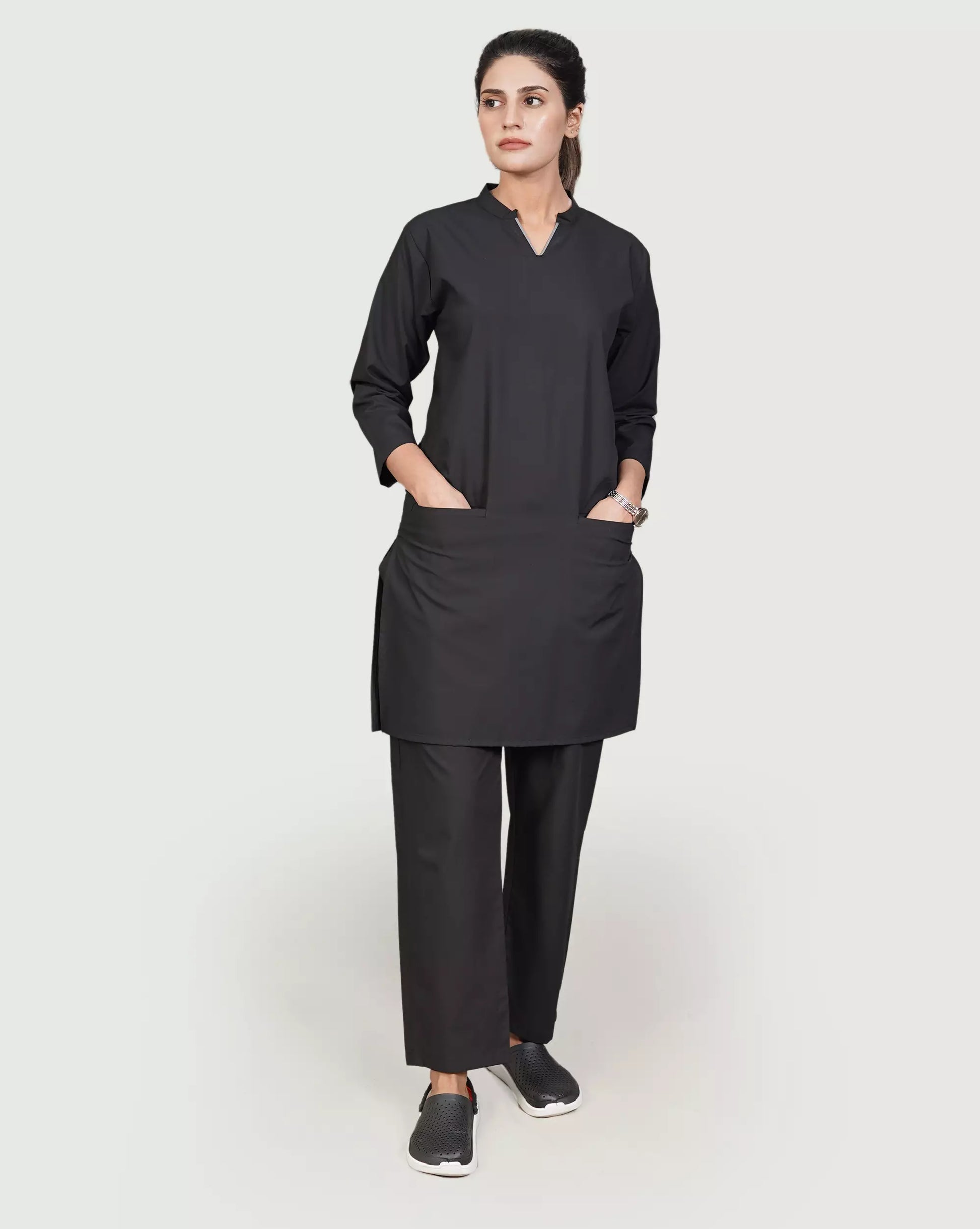 A woman standing, wearing a longtop with a V-neck design and two bone pockets, and matching trousers with two pockets. Both pieces are in a solid amber color.