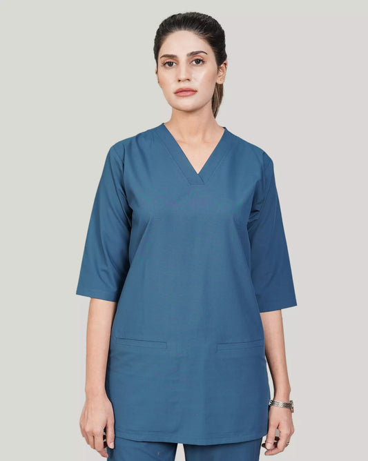 A woman wearing a green scrub set with a V-neckline and 3-quarter sleeves, featuring two bone pockets on the shirt and two pockets on the trousers. She is smiling and standing against a white background.