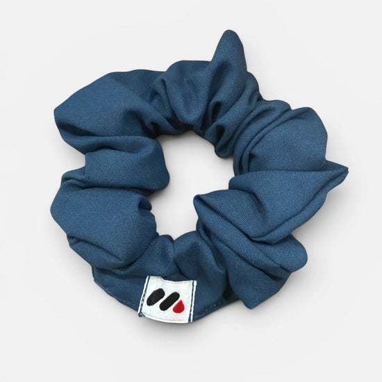 A dark blue fabric hair scrunchie with a small branded white tag featuring black and red abstract marks against a white background.