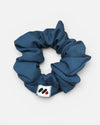A dark blue fabric hair scrunchie with a small branded white tag featuring black and red abstract marks against a white background.