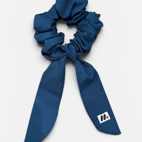 A dark blue fabric hair scrunchie with long tails and a small branded white tag featuring a black and red logo against a white background.