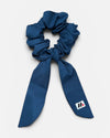 A dark blue fabric hair scrunchie with long tails and a small branded white tag featuring a black and red logo against a white background.