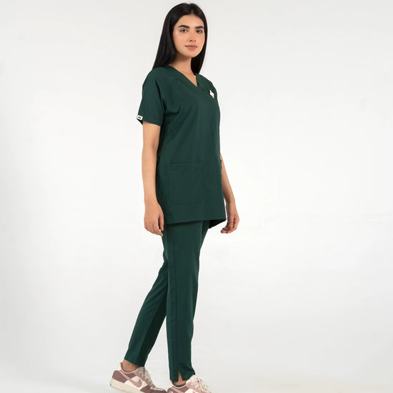 A healthcare professional modeling a dark green medical scrub set in a side profile view, wearing a short-sleeve V-top and matching pants with casual sneakers against a white background.