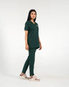 A healthcare professional modeling a dark green medical scrub set in a side profile view, wearing a short-sleeve V-top and matching pants with casual sneakers against a white background.