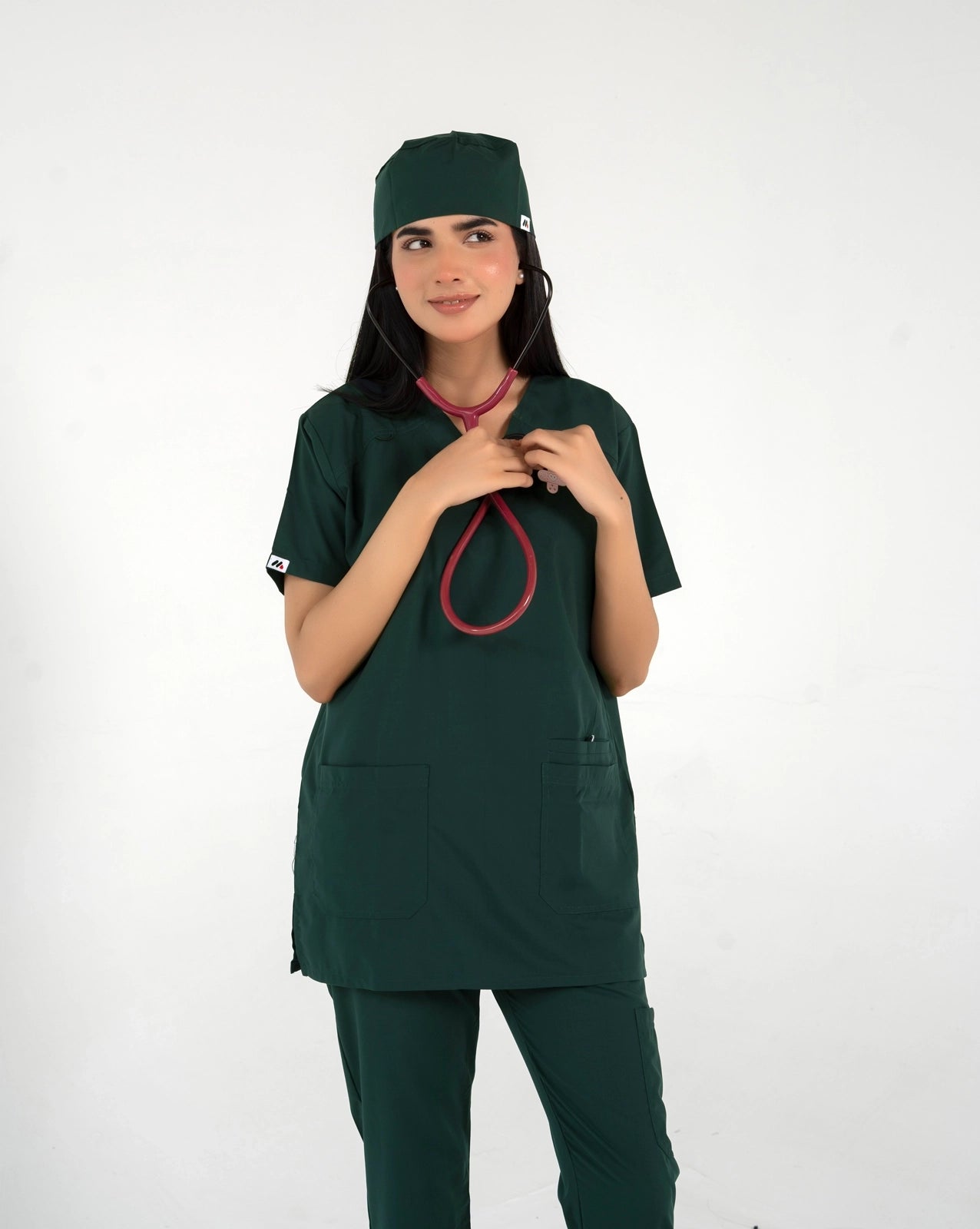 A healthcare professional wearing a dark green medical scrub set, matching scrub cap, and a red-tubed stethoscope around her neck against a white background.