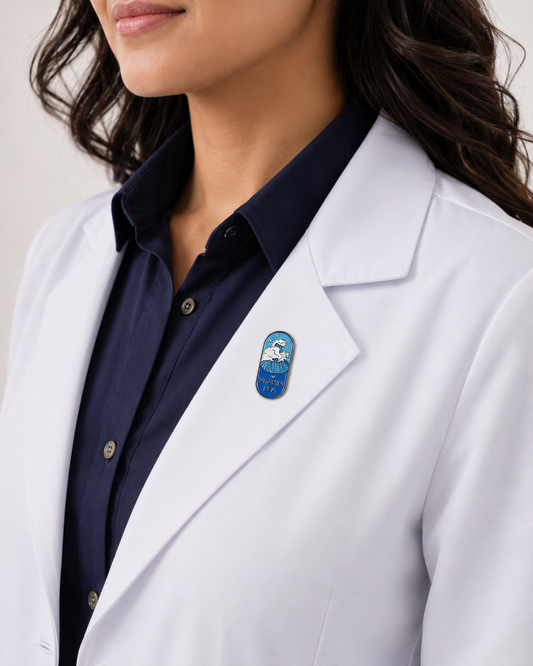 A close-up of a female healthcare professional wearing a white lab coat over a dark blue collared shirt with a blue and white capsule-shaped enamel lapel pin against a plain background.