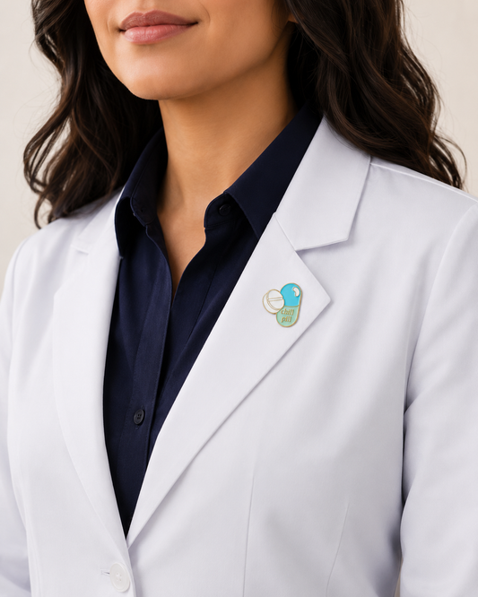 A close-up of a female healthcare professional wearing a white lab coat over a dark blue collared shirt with a capsule-shaped enamel lapel pin against a plain background.