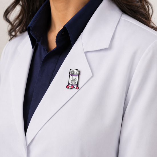 A close-up of a female healthcare professional wearing a white lab coat over a dark blue collared shirt with a prescription bottle enamel lapel pin against a plain background.