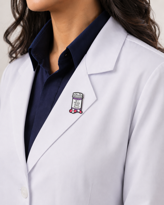 A close-up of a female healthcare professional wearing a white lab coat over a dark blue collared shirt with a prescription bottle enamel lapel pin against a plain background.