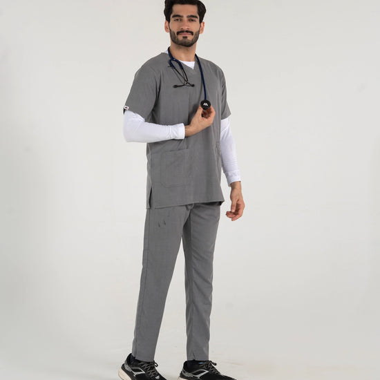 A male healthcare professional standing and modeling a full-body grey medical scrub set over a white long-sleeve undershirt, paired with black sneakers and a stethoscope against a white background.