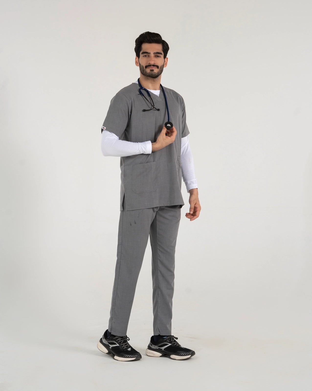 A male healthcare professional standing and modeling a full-body grey medical scrub set over a white long-sleeve undershirt, paired with black sneakers and a stethoscope against a white background.
