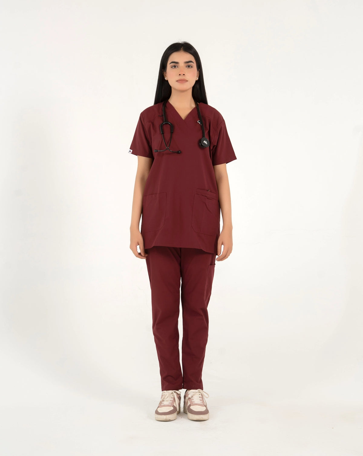 A healthcare professional standing and modeling a full-body maroon medical scrub set with a V-neck top, matching trousers, a stethoscope, and sneakers in a front view pose against a white background.