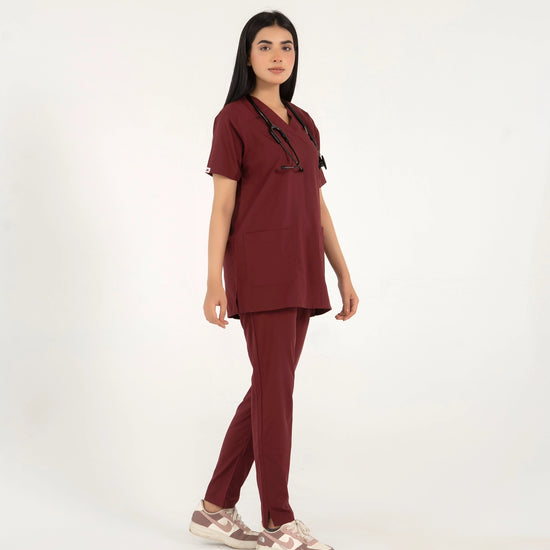 A healthcare professional standing and modeling a full-body maroon medical scrub set with a V-neck top, matching trousers, a stethoscope, and sneakers in a side view pose against a white background.
