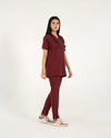 A healthcare professional standing and modeling a full-body maroon medical scrub set with a V-neck top, matching trousers, a stethoscope, and sneakers in a side view pose against a white background.