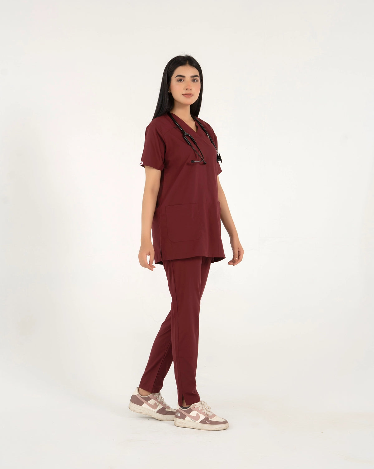 A healthcare professional standing and modeling a full-body maroon medical scrub set with a V-neck top, matching trousers, a stethoscope, and sneakers in a side view pose against a white background.