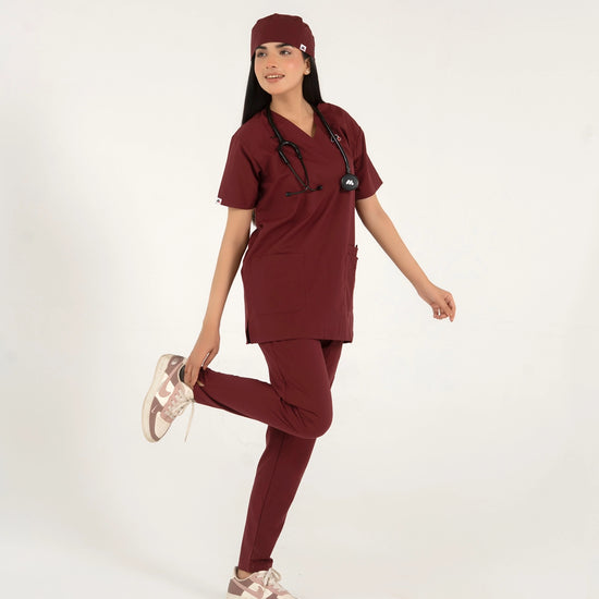 A healthcare professional standing and modeling a full-body maroon medical scrub set with a V-neck top, matching trousers, matching scrub cap, stethoscope, and sneakers while playfully lifting one foot against a white background.