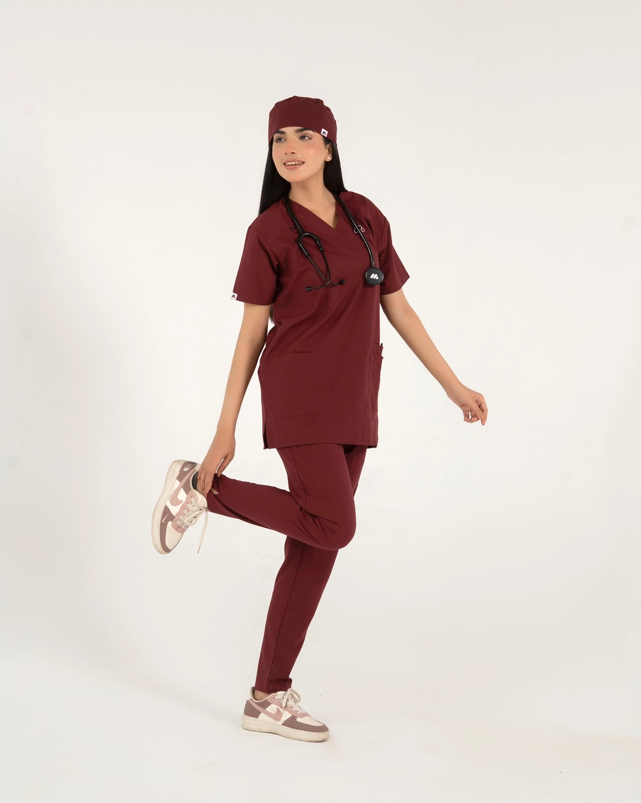 A healthcare professional standing and modeling a full-body maroon medical scrub set with a V-neck top, matching trousers, matching scrub cap, stethoscope, and sneakers while playfully lifting one foot against a white background.