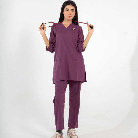 A healthcare professional standing and modeling a full-body purple medical scrub set while holding a stethoscope stretched across her shoulders against a white background.