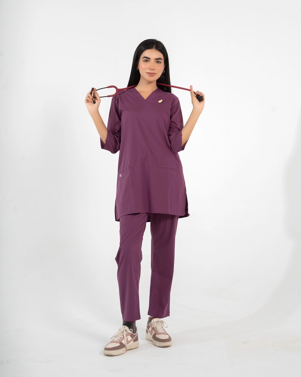 A healthcare professional standing and modeling a full-body purple medical scrub set while holding a stethoscope stretched across her shoulders against a white background.