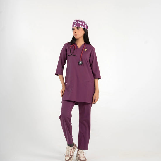 A healthcare professional standing and modeling a full-body purple medical scrub set, matching tooth-pattern scrub cap, stethoscope, and sneakers against a white background.