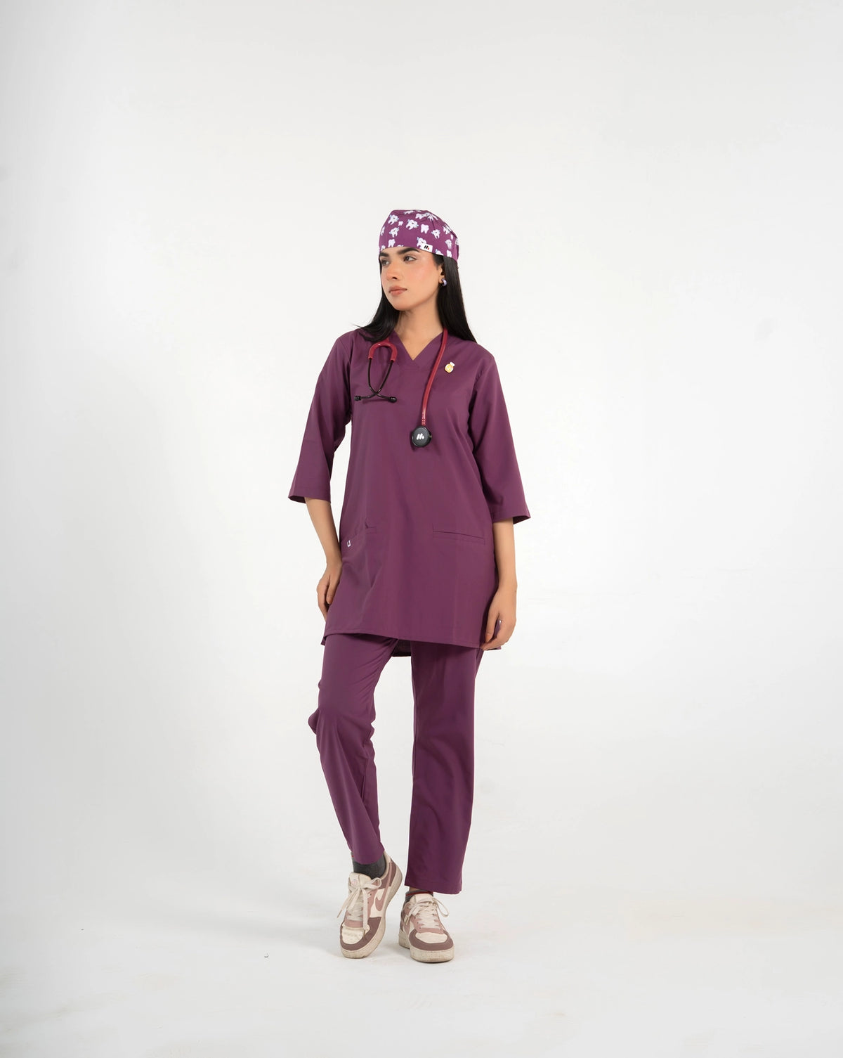 A healthcare professional standing and modeling a full-body purple medical scrub set, matching tooth-pattern scrub cap, stethoscope, and sneakers against a white background.