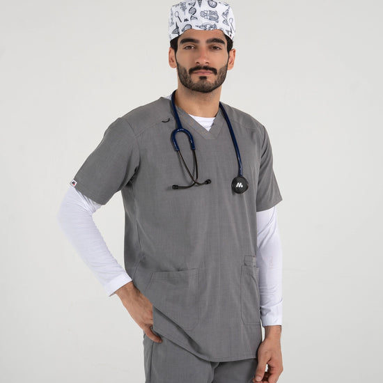 A male healthcare professional wearing a grey V-neck medical scrub set over a white long-sleeve undershirt, a stethoscope, and a white science-themed pattern scrub cap with one hand in his pocket against a white background.