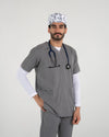 A male healthcare professional wearing a grey V-neck medical scrub set over a white long-sleeve undershirt, a stethoscope, and a white science-themed pattern scrub cap with one hand in his pocket against a white background.