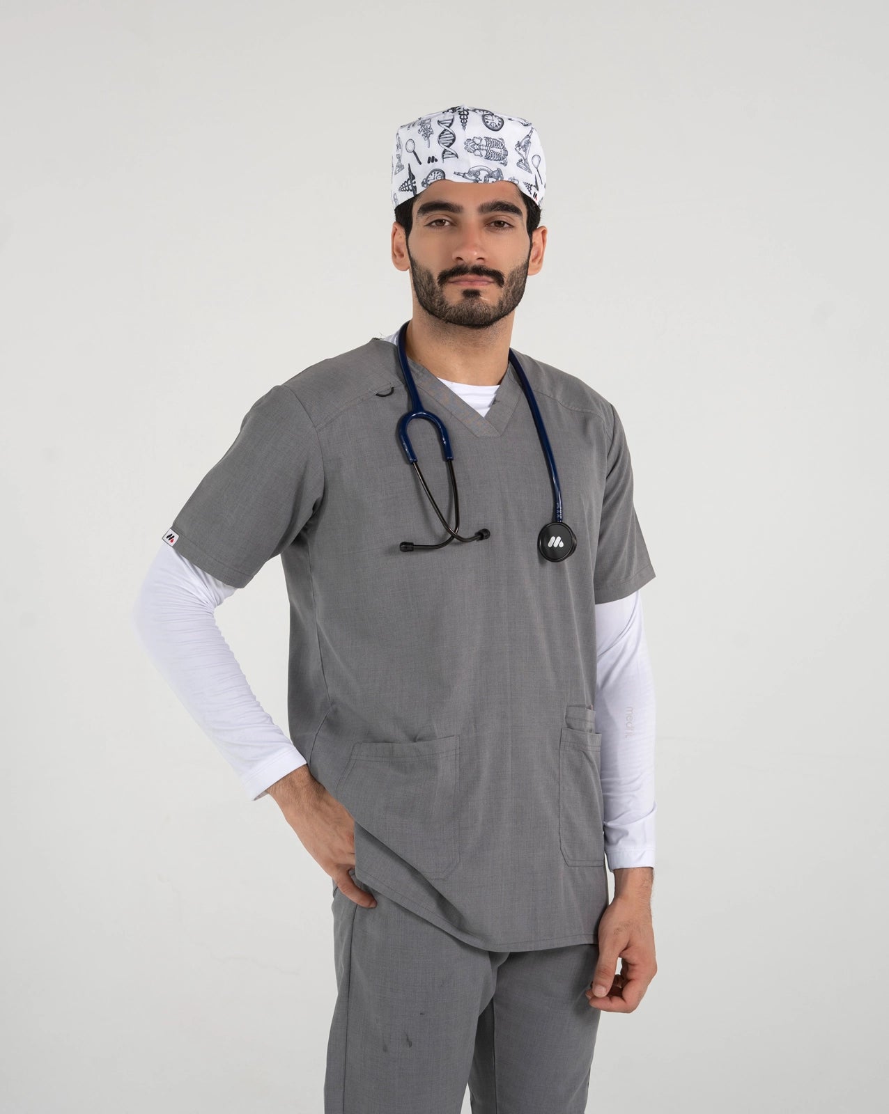A male healthcare professional wearing a grey V-neck medical scrub set over a white long-sleeve undershirt, a stethoscope, and a white science-themed pattern scrub cap with one hand in his pocket against a white background.