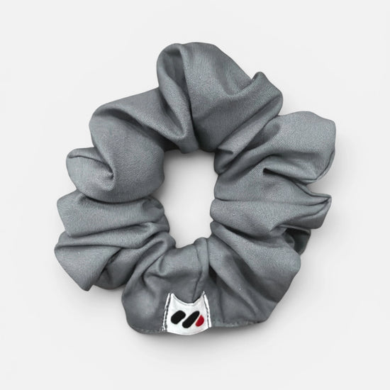 A grey fabric hair scrunchie with a small branded white tag featuring a black and red abstract mark against a white background.