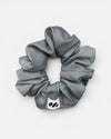 A grey fabric hair scrunchie with a small branded white tag featuring a black and red abstract mark against a white background.