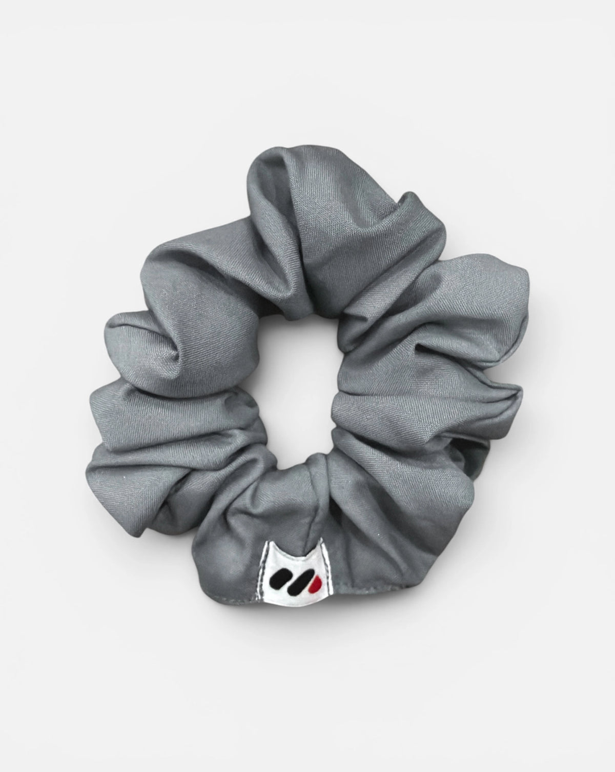 A grey fabric hair scrunchie with a small branded white tag featuring a black and red abstract mark against a white background.
