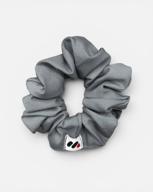 A grey fabric hair scrunchie with a small branded white tag featuring a black and red abstract mark against a white background.