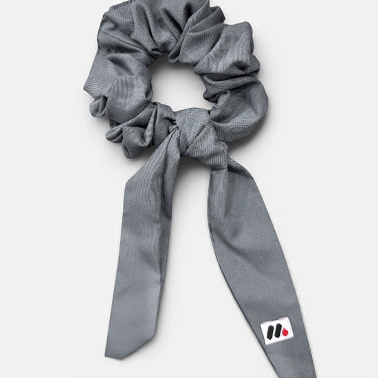A grey fabric hair scrunchie with long tails and a small branded white tag featuring a black and red logo against a white background.