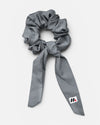 A grey fabric hair scrunchie with long tails and a small branded white tag featuring a black and red logo against a white background.