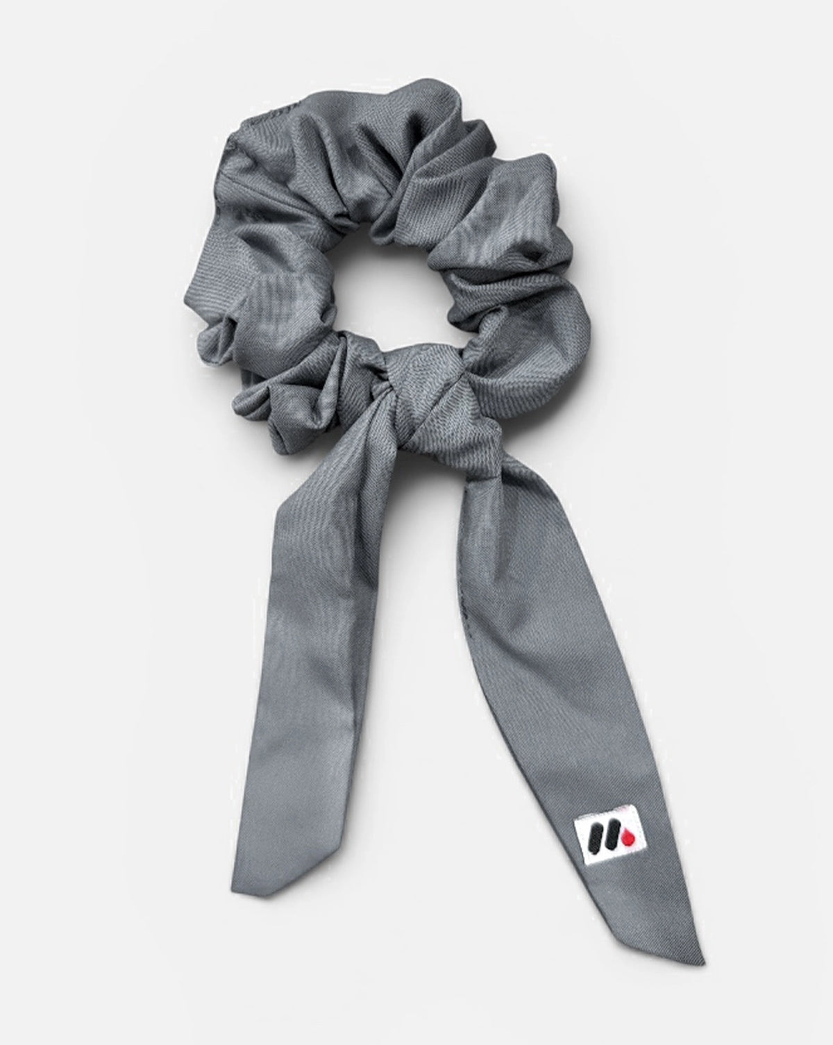 A grey fabric hair scrunchie with long tails and a small branded white tag featuring a black and red logo against a white background.