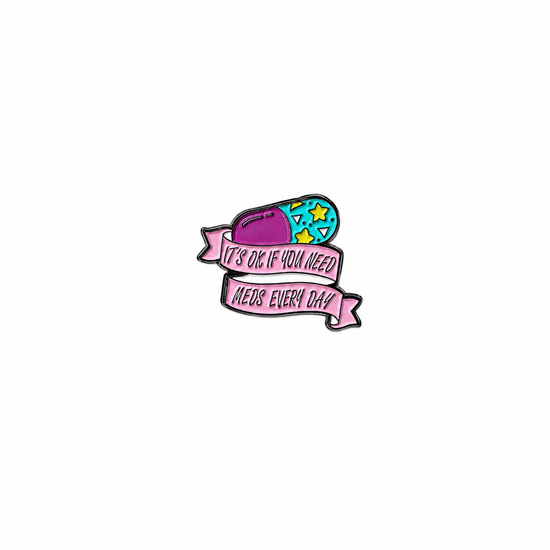 A colorful capsule enamel lapel pin paired with a pink banner featuring the text "IT'S OK IF YOU NEED MEDS EVERY DAY" against a white background.