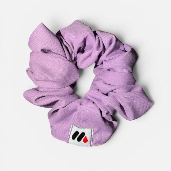 A lavender fabric hair scrunchie with a small branded white tag featuring a black and red abstract mark against a white background.